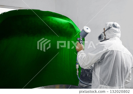 Automobile repairman painter in mask and protective workwear painting car hood to green color in paint chamber. Automobile repairman painter in mask and protective workwear painting car hood to green color in paint chamber. 132020802