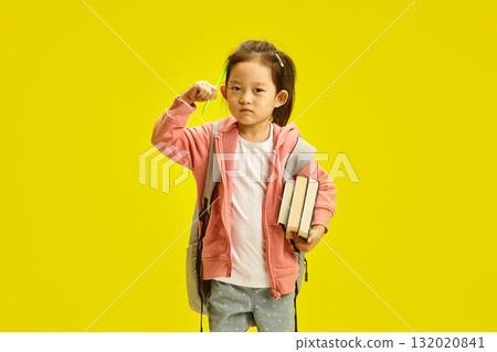 Contemplative Asian first grader girl dressed in pink hoodie holds her school books and pen near temple standing over yellow isolated. Distance education for preschool concept. 132020841