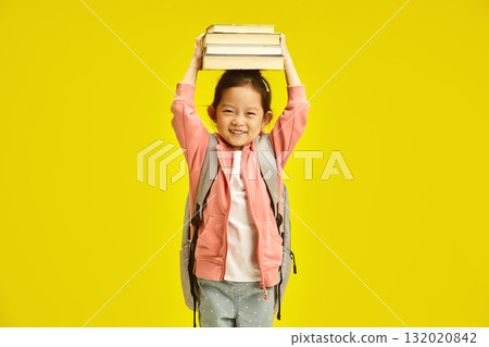Cute chinese schoolgirl holding books on head over yellow isolated background. First grader kid girl preparing to the school. Cute chinese schoolgirl holding books on head over yellow isolated background. First grader kid girl preparing to the school. 132020842