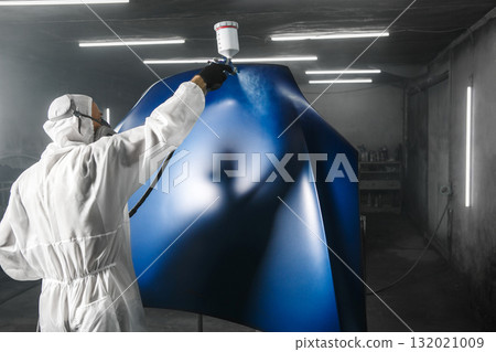 Professional bodywork, male car painter in protective mask and workwear apply few layer paint from spray gun to automobile body parts in garage booth.  132021009
