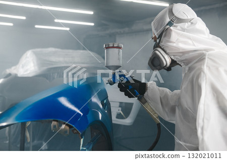 Worker in protective mask and jumpsuit overall spraying base coat of paint on car body elements in painted chamber using professional airbrush. 132021011