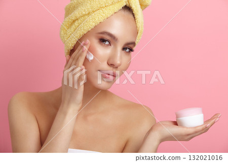 Seductive female model applies cream on cheek at pink wall 132021016