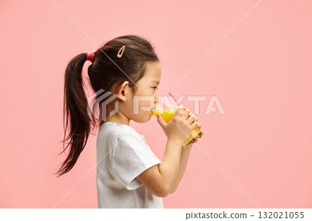 Profile Portrait of Sweet Ponytail Little Girl Drinking Orange Juice From Round Glass and Looking to Camera Standing Over Pink Isolated Profile Portrait of Sweet Ponytail Little Girl Drinking Orange Juice From Round Glass and Looking to Camera Standing Over Pink Isolated 132021055