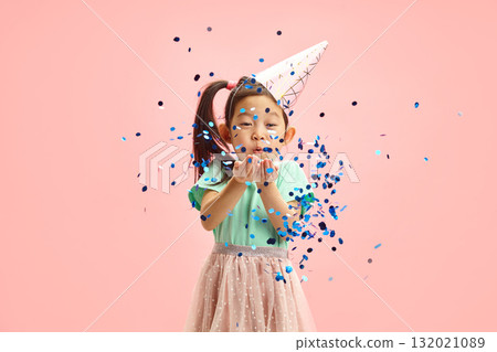 Waist up portrait of Sweet Asian Child Girl Blowing Blue Glitter to Camera While Standing Against Pink Isolated Background  132021089