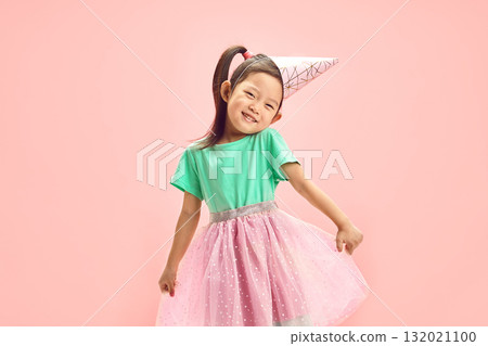 Cheerful Birthday Asian Child Girl in Festive Clothes, Having Party Hat on Head, Cute Raising Shoulders, Funny Straightens her full Skirt to the Sides Standing Over Pink Isolated Backgound. Cheerful Birthday Asian Child Girl in Festive Clothes, Having Party Hat on Head, Cute Raising Shoulders, Funny Straightens her full Skirt to the Sides Standing Over Pink Isolated Backgound. 132021100