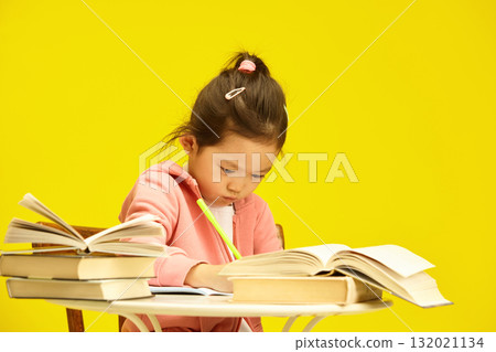 Studious Asian preschooler child girl deep in study amidst a pile of books sitting at table over sunny yellow isolated background, writing somethnig in copybook Studious Asian preschooler child girl deep in study amidst a pile of books sitting at table over sunny yellow isolated background, writing somethnig in copybook 132021134