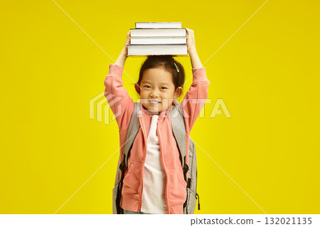 Sweet first grader asian ethnicity schoolgirl holding books above head, cheerful smiling standing with backpack on her shoulders over yellow isolated background. Sweet first grader asian ethnicity schoolgirl holding books above head, cheerful smiling standing with backpack on her shoulders over yellow isolated background. 132021135