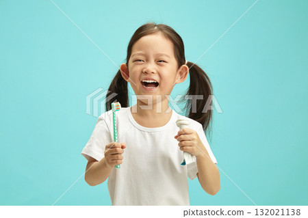 Cheerful little asian ethnicity girl holding squeezed toothpaste tube and toothbrush, fun laughing, wearing in white t shirt, standing over blue isolated background. Cheerful little asian ethnicity girl holding squeezed toothpaste tube and toothbrush, fun laughing, wearing in white t shirt, standing over blue isolated background. 132021138