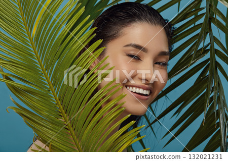 Playful girl peeks out from behind palm leaves, smiles. 132021231
