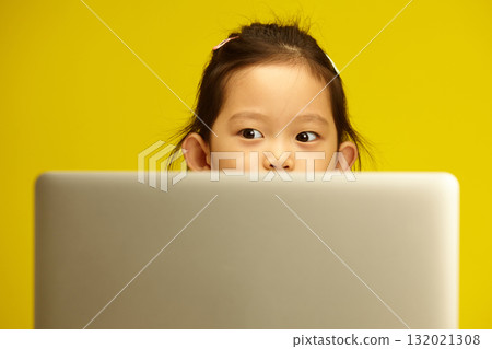 Pensive Asian Preschool Girl with a big opened eyes Engaged in Distance Learning via Laptop sitting against yellow isolated background Pensive Asian Preschool Girl with a big opened eyes Engaged in Distance Learning via Laptop sitting against yellow isolated background 132021308