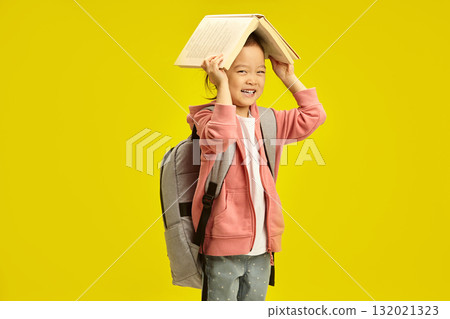 Little japanese smiling schoolgirl with book and backpack on her shoulders posing against yellow background. Asian ethnicity cheerful child girl holds opening book above head. Little japanese smiling schoolgirl with book and backpack on her shoulders posing against yellow background. Asian ethnicity cheerful child girl holds opening book above head. 132021323