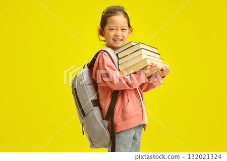 Cheerful little korean first grader girl in casual clothes with many books for studying at school and backpack on shoulders isolated over yellow background 132021324