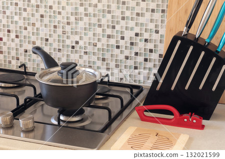 Saucepan with lid on gas stove and knife in stand at home. Cooking at home. Mosaic tiles on kitchen worktop. Saucepan with lid on gas stove and knife in stand at home. Cooking at home. Mosaic tiles on kitchen worktop. 132021599