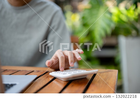 Close up of a hand using a white calculator while sitting at wooden table. Study or working outdoor. 132022218