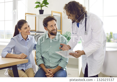Smiling happy young couple sitting with a male family doctor therapist in medical clinic. 132025537