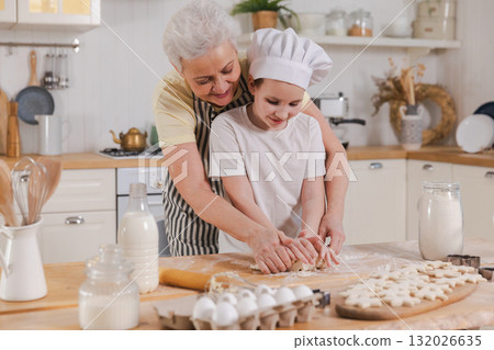 Happy family in kitchen. Grandmother and granddaughter child cook in kitchen together. Grandma teaching kid girl knead dough bake cookies. Household teamwork helping family generations concept Happy family in kitchen. Grandmother and granddaughter child cook in kitchen together. Grandma teaching kid girl knead dough bake cookies. Household teamwork helping family generations concept 132026635