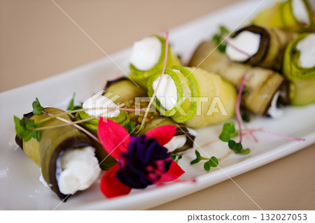 Female chef hands preparing healthy zucchini and eggplant rolls stuffed creamy feta cheese and roasted seeds on white plate surrounded by fresh vegetables in a bright modern kitchen restaurant scene. Female chef hands preparing healthy zucchini and eggplant rolls stuffed creamy feta cheese and roasted seeds on white plate surrounded by fresh vegetables in a bright modern kitchen restaurant scene. 132027053