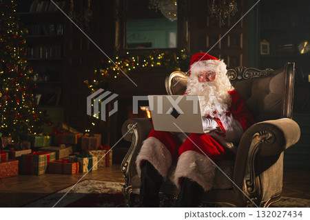 Merry Christmas. Santa Claus with laptop reading letters from children typing buying presents gifts online. Santa with computer on Christmas eve. Mail gifts of Santa Claus. Winter Holiday Celebrations 132027434