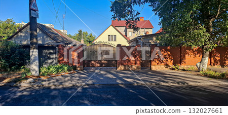 Street view of a house with a red roof and brick fence. House with a prominent red roof and beige facade set behind a brick fence.  132027661