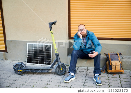 Male person waits while charging electric scooter with photovoltaic solar panel in urban settings. Integration of solar power as sustainable energy source, promoting eco-friendly urban transportation. 132028170