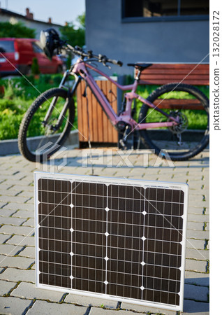 Pink electric mountain bike parked next to wooden bench, connected to solar panel for charging on a blurred background. Bike rests on paved area surrounded by greenery and grass. 132028172