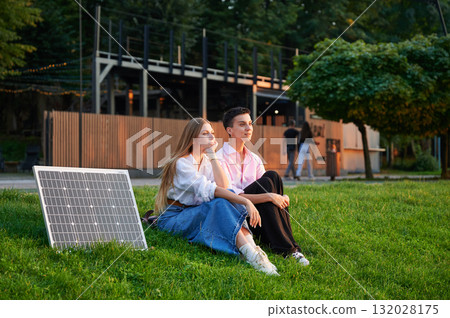 Young women using solar panel for charging, sit on grassy lawn, smiling and talking, with solar panel placed nearby. Girls dressed casually, enjoying sunny, relaxed moment outdoors. 132028175