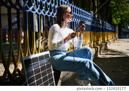 Happy woman connecting smartphone to photovoltaic solar panel. Integration of sustainable renewable energy into everyday life, demonstrating practical use of solar power for charging devices. Happy woman connecting smartphone to photovoltaic solar panel. Integration of sustainable renewable energy into everyday life, demonstrating practical use of solar power for charging devices. 132028177