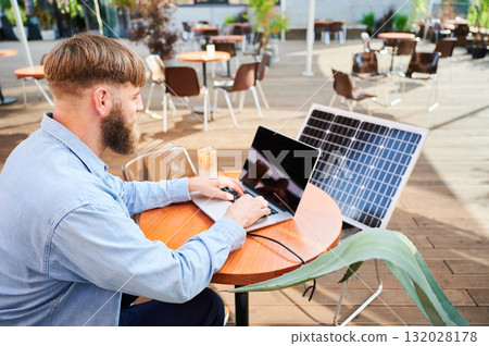 Man works on laptop at outdoor round table, with glass of iced coffee. Solar panel nearby, emphasizing sustainable, modern eco-friendly workspace that combines technology and sustainability. 132028178