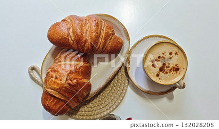 Two golden croissants served on a ceramic plate with a cup of cappuccino sprinkled with cinnamon. Concept of cozy breakfast, comfort, and slow morning indulgence at home or cafe 132028208