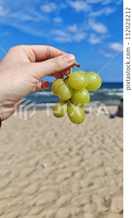 A hand holding a bunch of green grapes against a bright blue sky and sandy beach background. Concept of summer freshness, healthy eating, and carefree seaside relaxation A hand holding a bunch of green grapes against a bright blue sky and sandy beach background. Concept of summer freshness, healthy eating, and carefree seaside relaxation 132028212