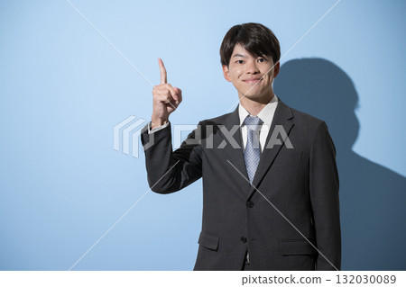 A young businessman in a suit, likely a new graduate, is pointing and looking at the camera. Image of job hunting, light blue background 132030089