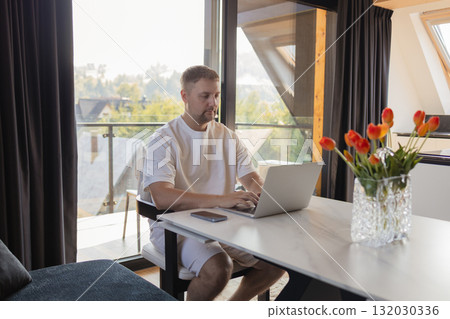 Man working on laptop at home office with flowers on table. . Modern workspace with natural light, tulips on the table and cozy atmosphere. Man working on laptop at home office with flowers on table. . Modern workspace with natural light, tulips on the table and cozy atmosphere. 132030336