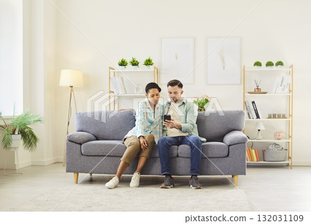Happy couple sitting on sofa in living room at home and using smartphone together browsing apps. Happy couple sitting on sofa in living room at home and using smartphone together browsing apps. 132031109