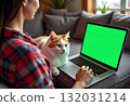 An attractive young woman is working on a laptop while sitting cross-legged in a comfortable chair at home with a funny assistant cat on her legs. 132031214