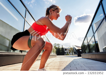 Athlete woman in bright sports bra squats outdoors during sunny workout session between glass buildings 132031952
