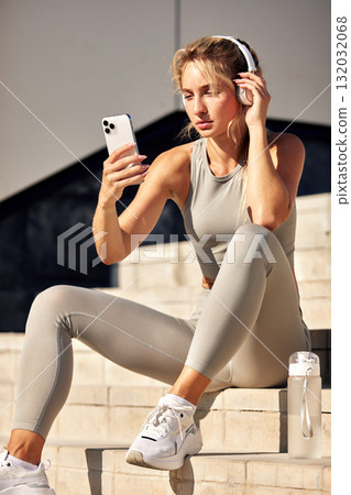 Athletic woman with headphones checking her phone on outdoor steps, ready for a workout Athletic woman with headphones checking her phone on outdoor steps, ready for a workout 132032068