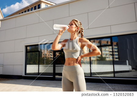 Athlete drinks protein shake after outdoor workout beside modern building, fitness training scene 132032083