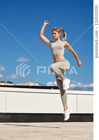 Strong athletic woman jumps in athletic wear on rooftop under clear blue sky during a sunny day 132032091