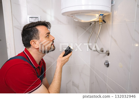 Plumber inspecting water heater with flashlight in bathroom Plumber inspecting water heater with flashlight in bathroom 132032946