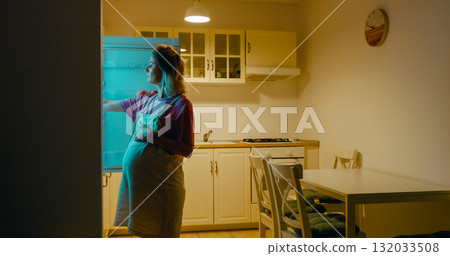 Pregnant woman opening refrigerator and eating cake at home. 132033508