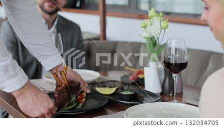 Waiter brings order rack of lamb for couples in restaurant. Couple sitting by table in a cafe. Customer service. 132034705