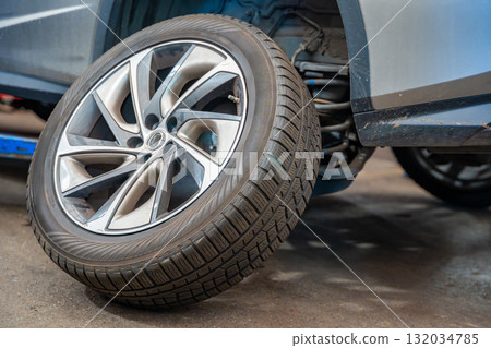 Prepared car wheel lying next to the vehicle before final installation. Seasonal tire change process during winter preparation at auto service workshop. 132034785