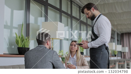 Waiter takes an order from a young couple in restaurant. Cafe worker taking note, writing order. Customer service. 132034798