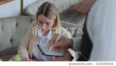 Waiter talking to woman sitting at table placing order in restaurant. Waiter takes an order on a tablet. Waiter talking to woman sitting at table placing order in restaurant. Waiter takes an order on a tablet. 132034834