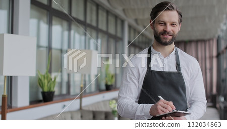 Young waiter man writes down an order in a notebook in a restaurant. Portrait of a young waiter in a white shirt and apron smiling in a restaurant. 132034863