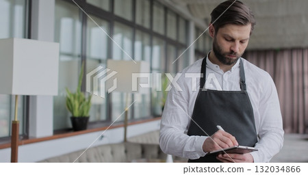 Young waiter man writes down an order in a notebook in a restaurant. Portrait of a young waiter in a white shirt and apron smiling in a restaurant. 132034866