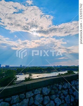 Scenic river view with sun rays through clouds. Panoramic view over a river and distant city skyline with dramatic sun rays breaking through the clouds above a stone wall. Scenic river view with sun rays through clouds. Panoramic view over a river and distant city skyline with dramatic sun rays breaking through the clouds above a stone wall. 132035742