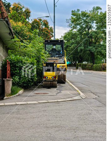 Two yellow road rollers parked on the street. Large and small yellow road rollers parked by the roadside near green trees. Construction machinery and heavy equipment outdoors. Two yellow road rollers parked on the street. Large and small yellow road rollers parked by the roadside near green trees. Construction machinery and heavy equipment outdoors. 132035807