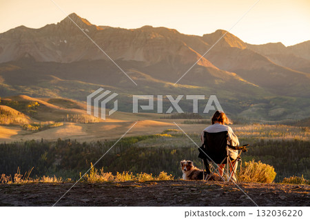 Female traveler sitting on a camping chair with her dog watching the mountain view during sunset. Female traveler sitting on a camping chair with her dog watching the mountain view during sunset. 132036220