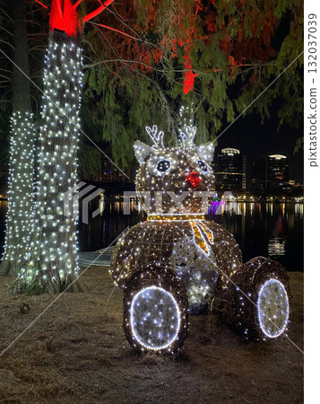 A reindeer-shaped light sculpture glows under trees beside the city lake. The reflection of lights creates a peaceful and festive tropical night view. A reindeer-shaped light sculpture glows under trees beside the city lake. The reflection of lights creates a peaceful and festive tropical night view. 132037039